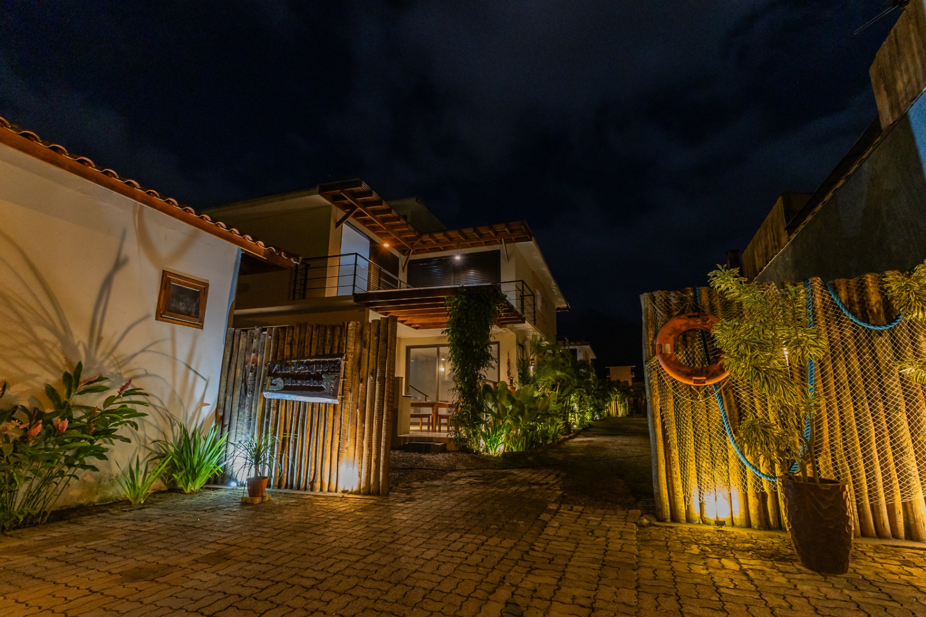Entrada iluminada da Vila Paquetá à noite, com portal de bambu e jardim