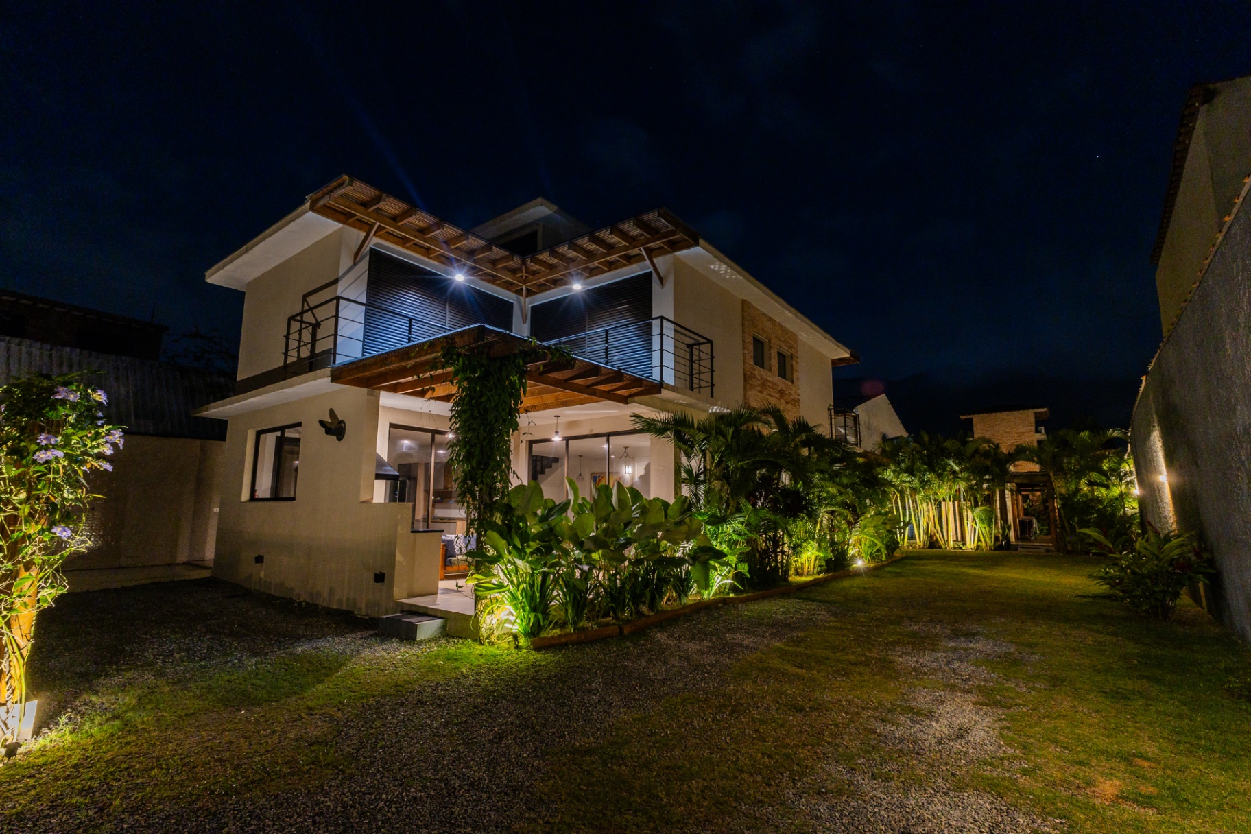 Fachada frontal iluminada à noite com pergolado e jardim tropical